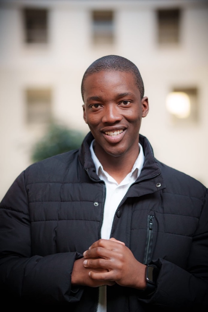 John Mokwena smiling with his hands clasped, wearing a black padded jacket over a white shirt